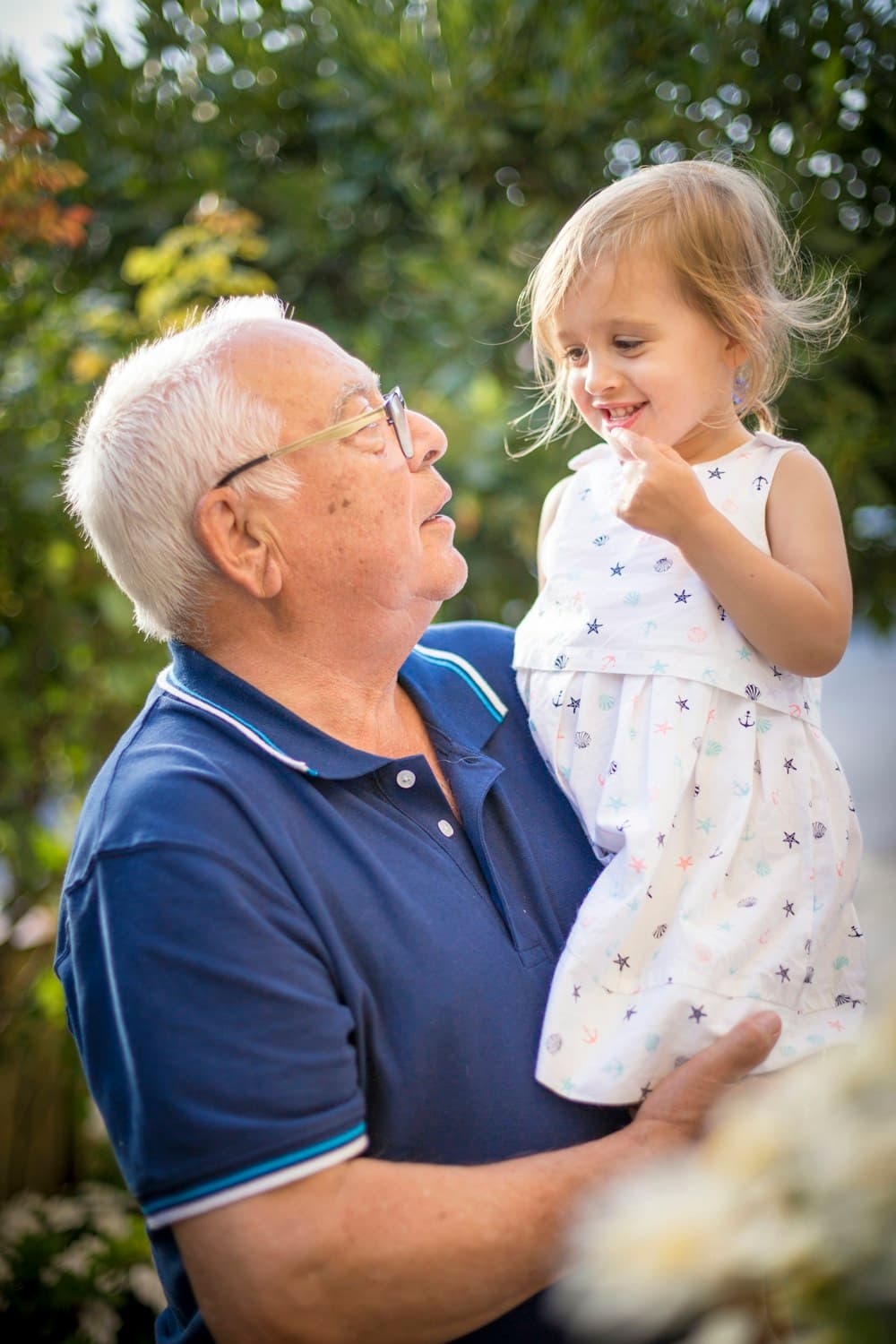 In einem Garten hält ein weißhaariger Großvater mit Brille ein kleines Mädchen im Arm; die beiden sehen sich an, das Mädchen lächelt.